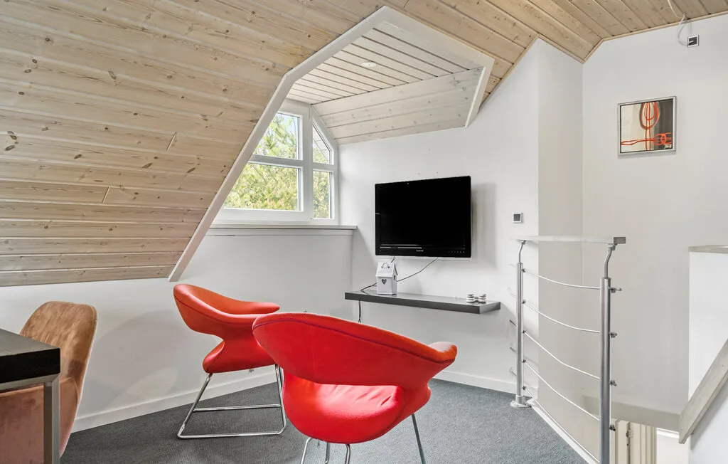 Una luminosa sala bajo el techo tiene asientos rojos, un televisor en la pared y techo inclinado de madera.