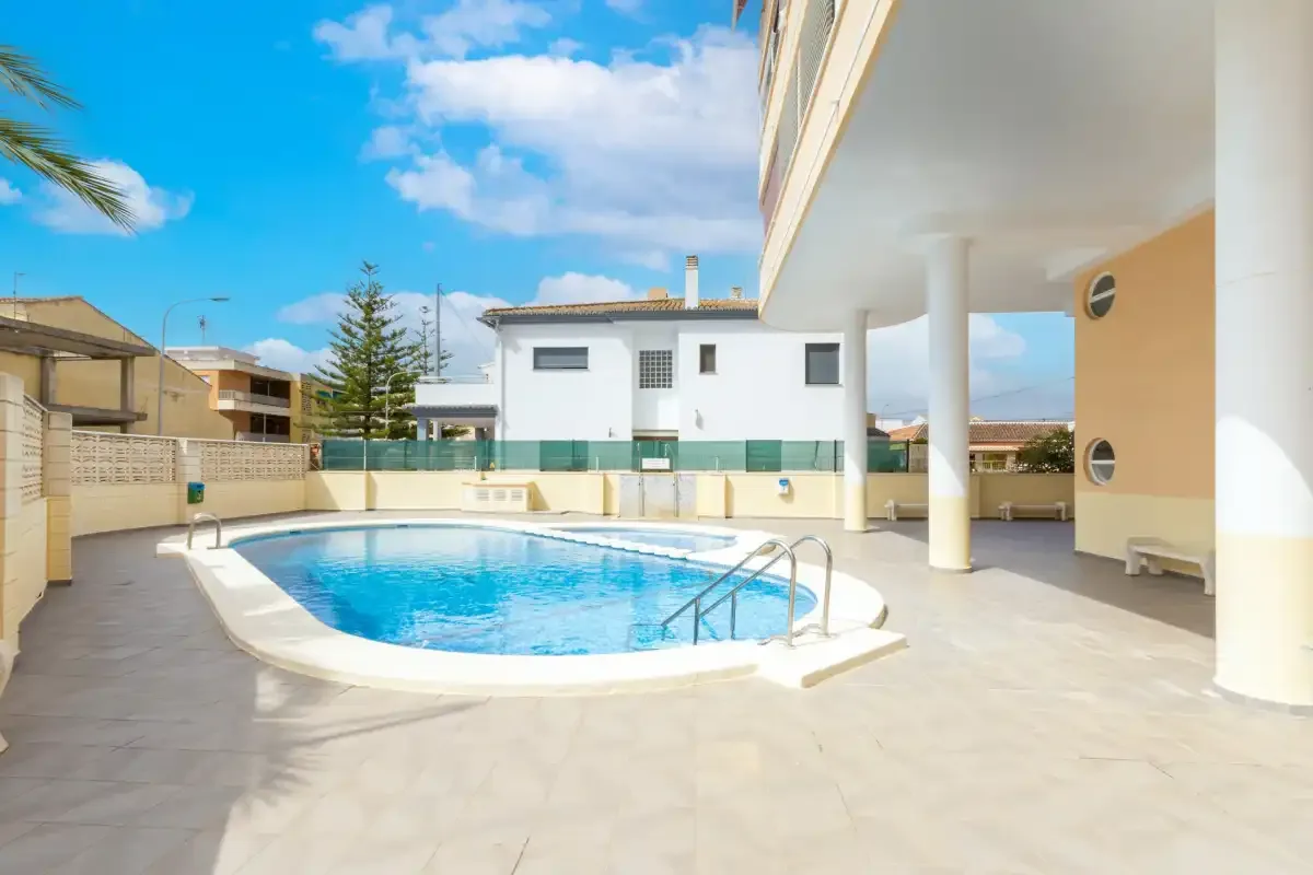 An outdoor swimming pool sits beside a covered patio and surrounding buildings.