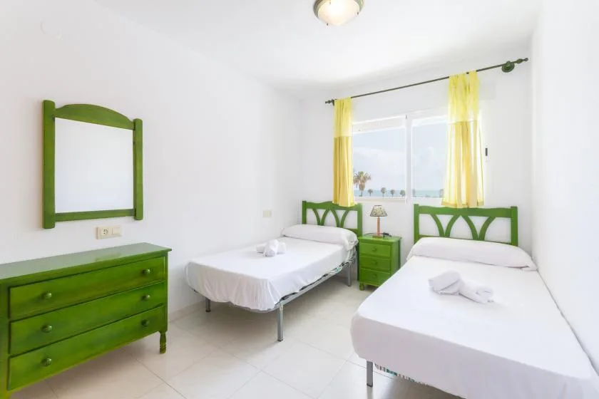 A bright bedroom with two beds and green furniture beneath a window.