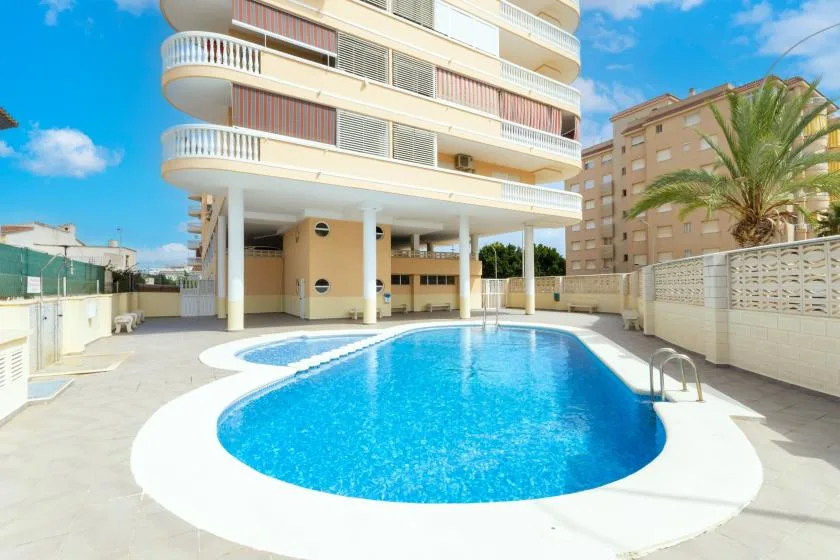 An outdoor swimming pool sits beside a multi-story building under a blue sky.