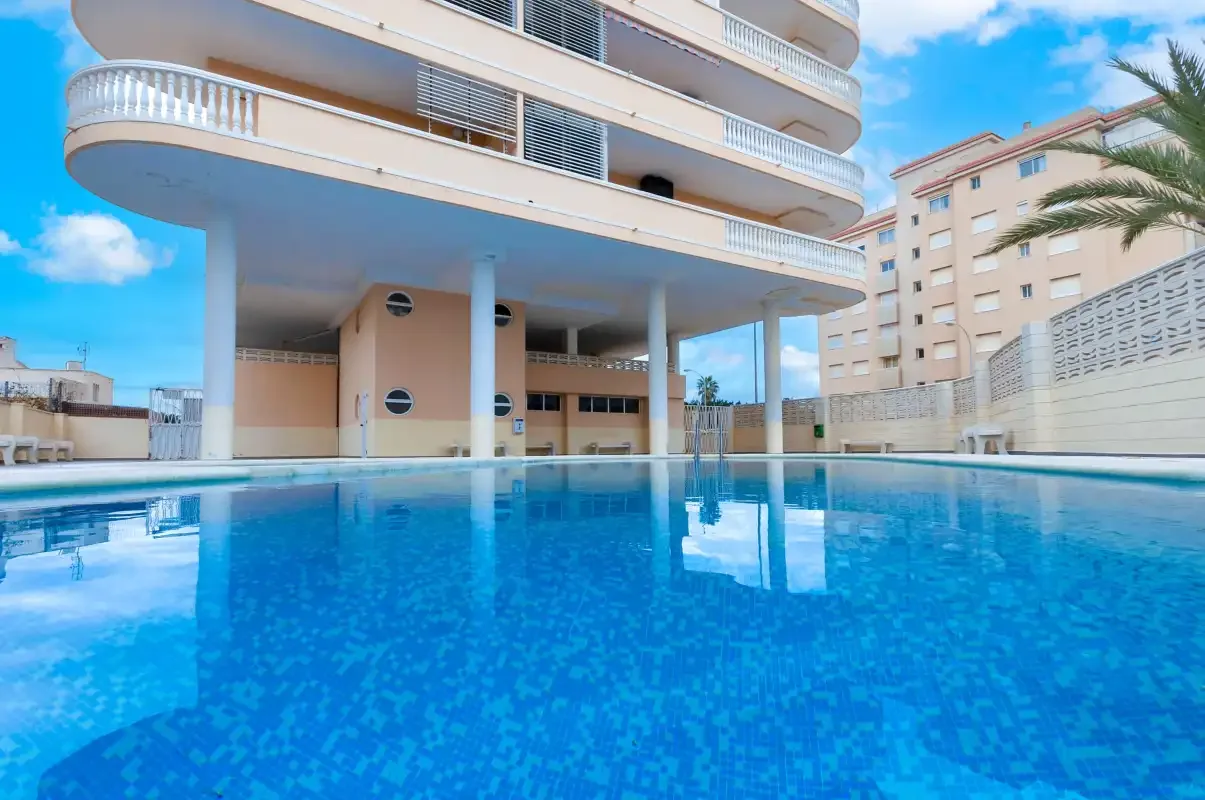 A swimming pool sits beside a curved apartment building under a bright blue sky.