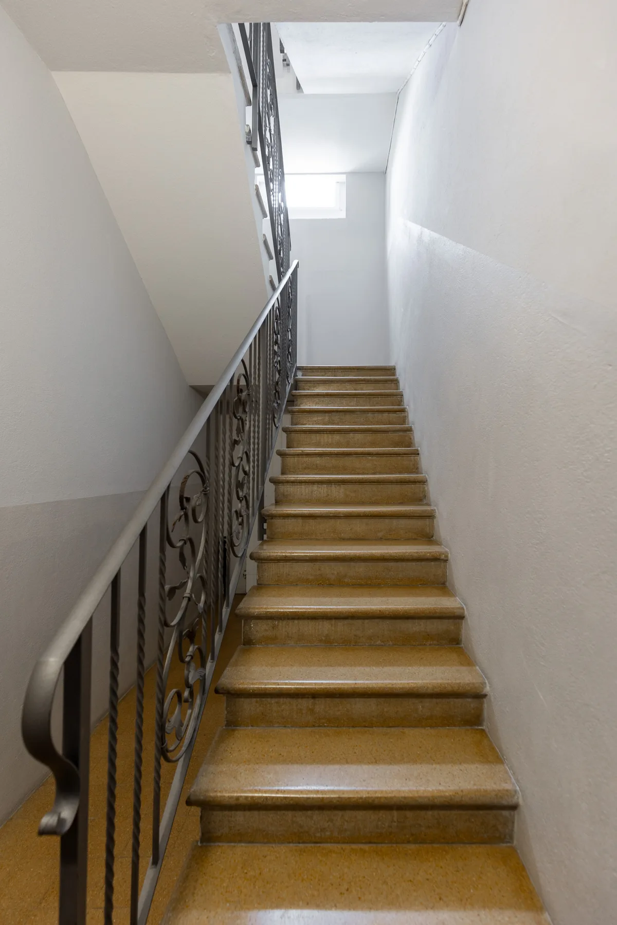 Una escalera interior estrecha con peldaños de madera y una barandilla metálica decorativa.