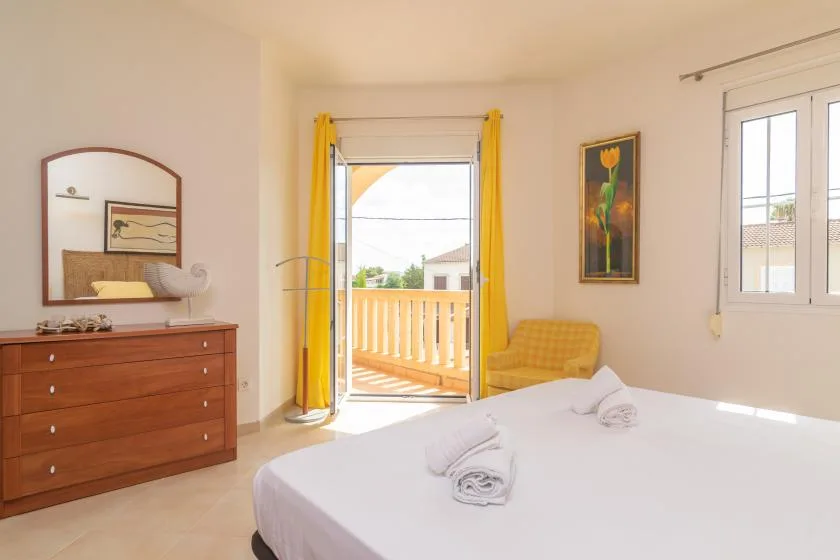 Un dormitorio luminoso con una cama, una cómoda de madera y puertas de vidrio que dan a un balcón.