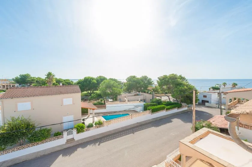 Una calle residencial soleada con casas bajas, árboles y una vista parcial del mar.