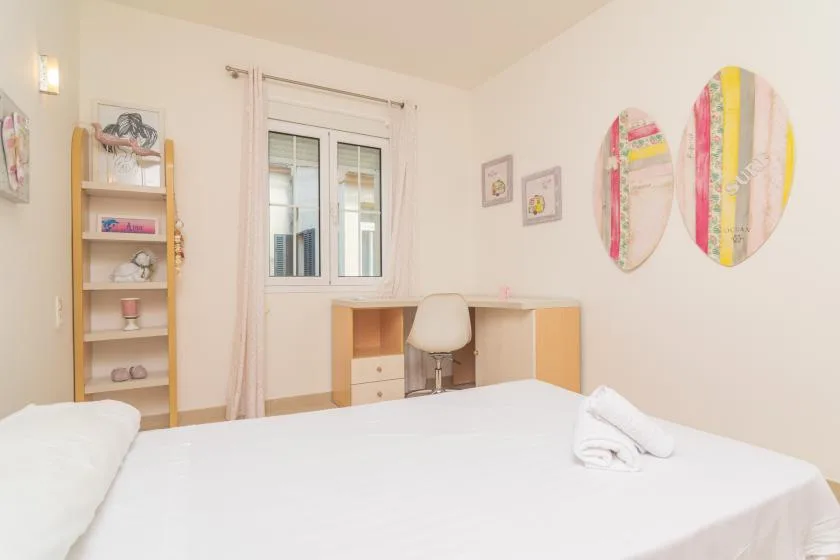 Un dormitorio luminoso con una cama blanca, un escritorio y adornos de colores en la pared.