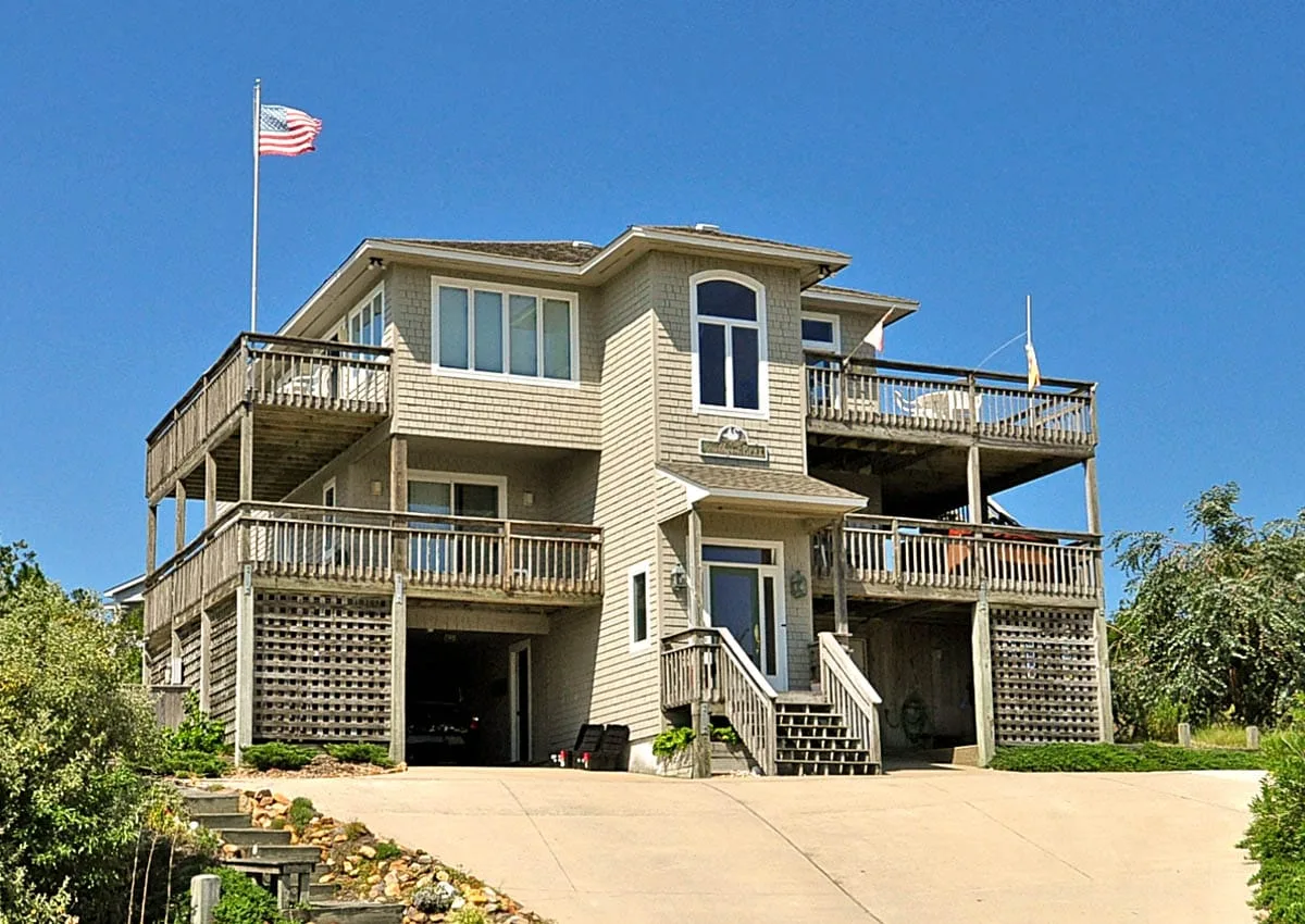 A large raised house with wooden decks, a central entrance, and a broad driveway.