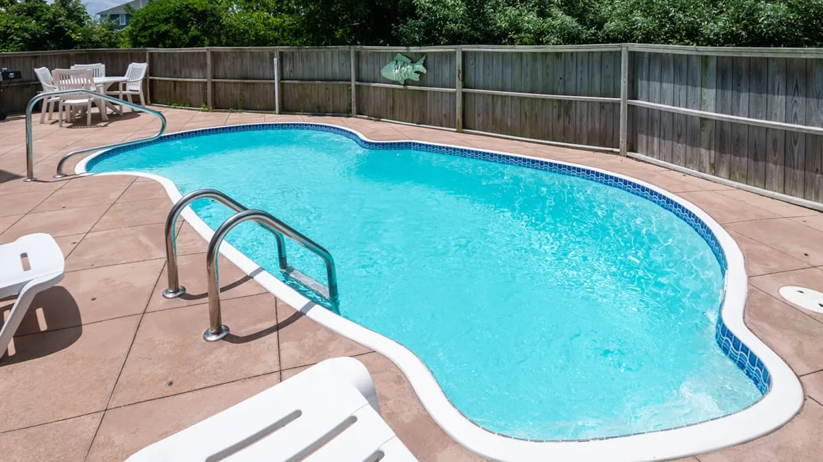 An outdoor swimming pool is surrounded by a paved deck and wooden fence.