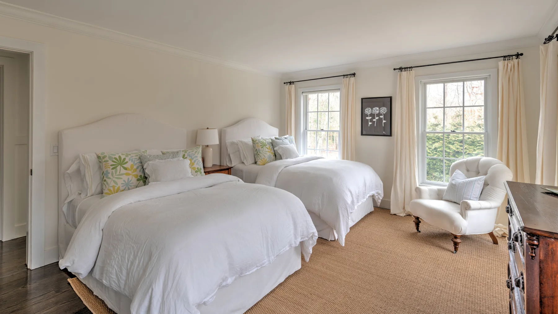 Un dormitorio luminoso con dos camas y un sillón junto a las ventanas.