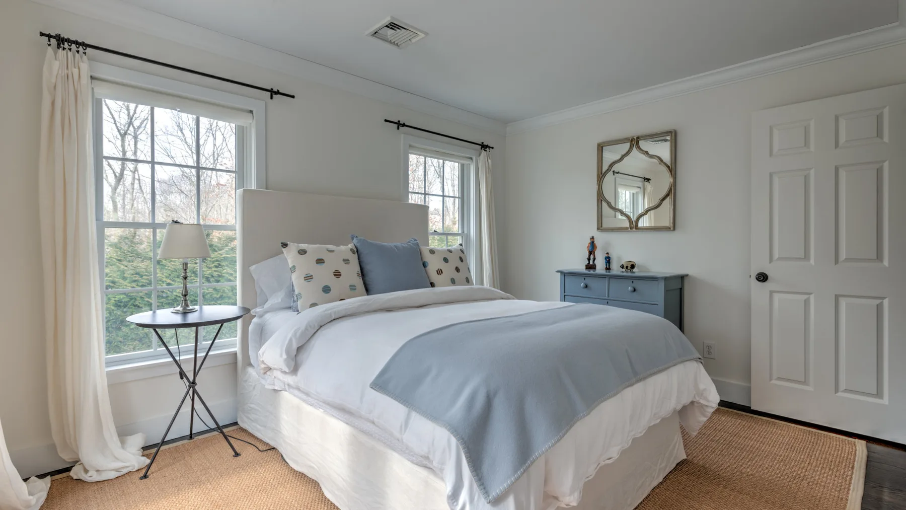 Un dormitorio luminoso con una cama tendida y ventanas con vista a los árboles.