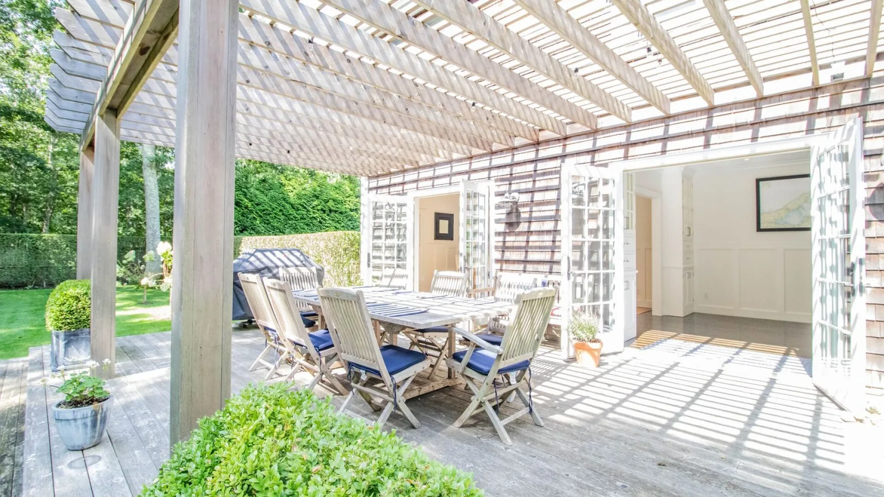 Comedor al aire libre bajo una pérgola de listones, con mesa y asientos.