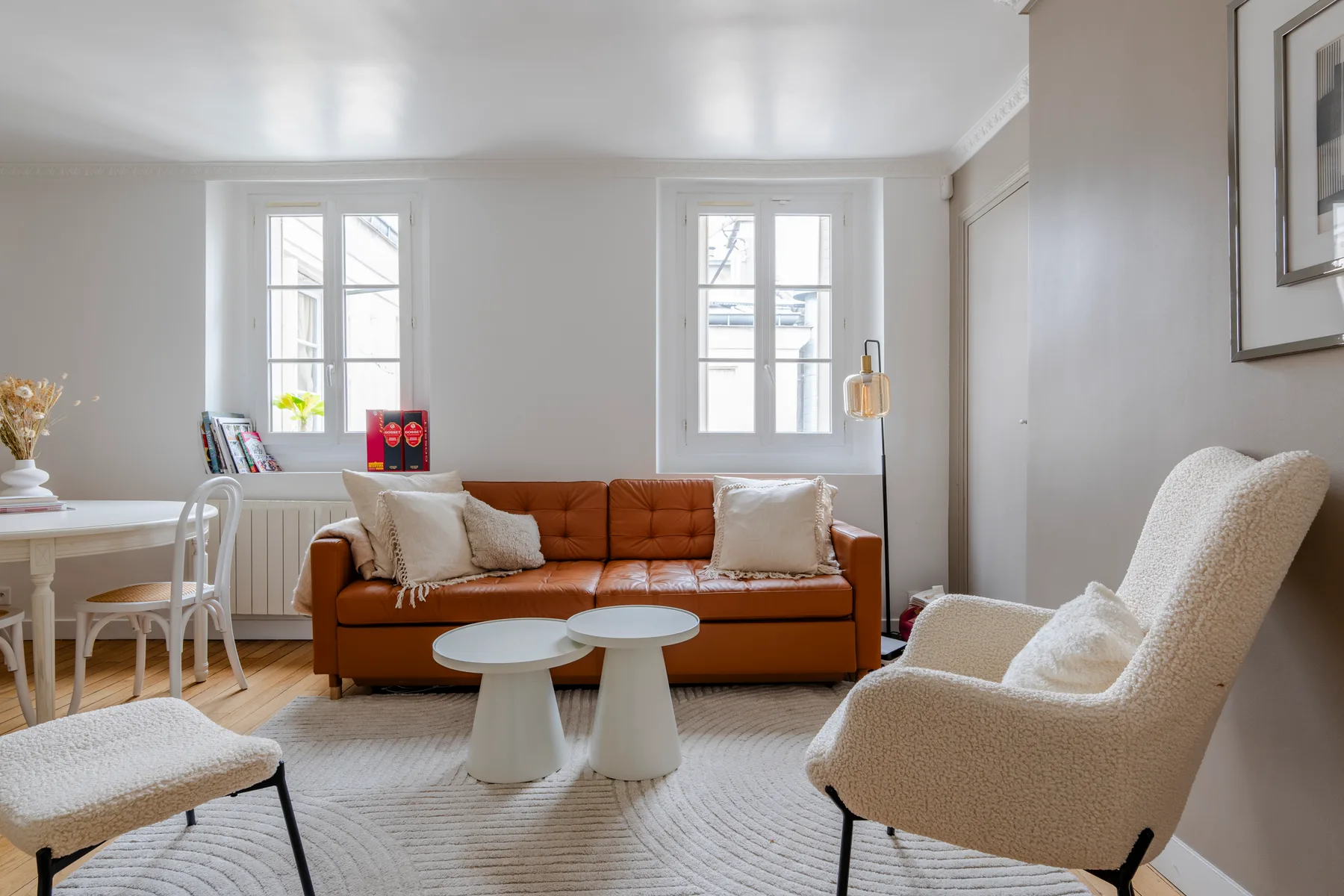 Una sala luminosa con un sofá naranja, asientos blancos y dos ventanas.