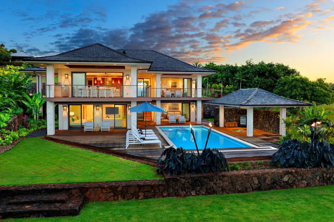 Una casa grande da a una piscina y un césped verde al atardecer.