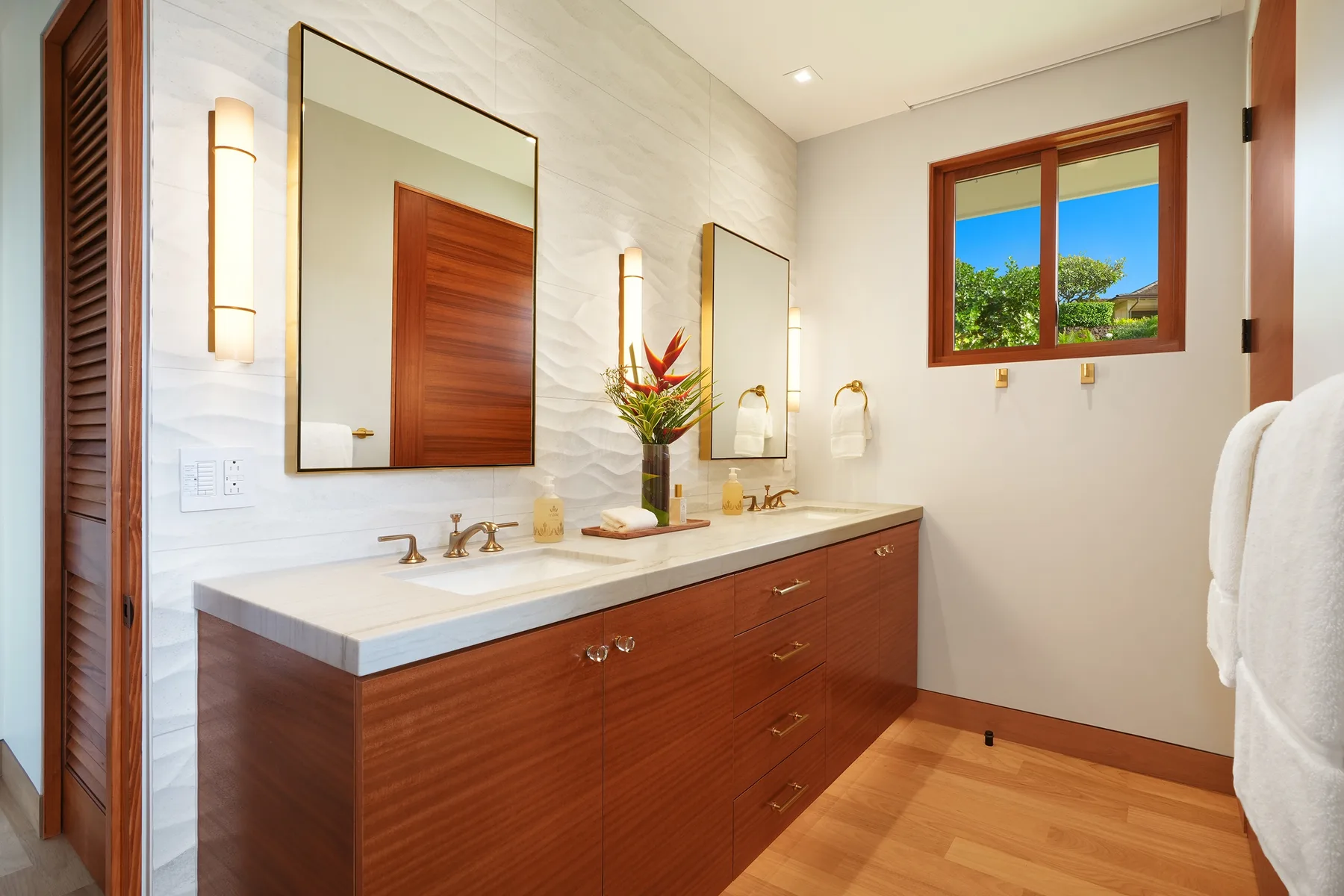 Un baño luminoso con un lavabo largo, espejos y una ventana con vista a la vegetación.
