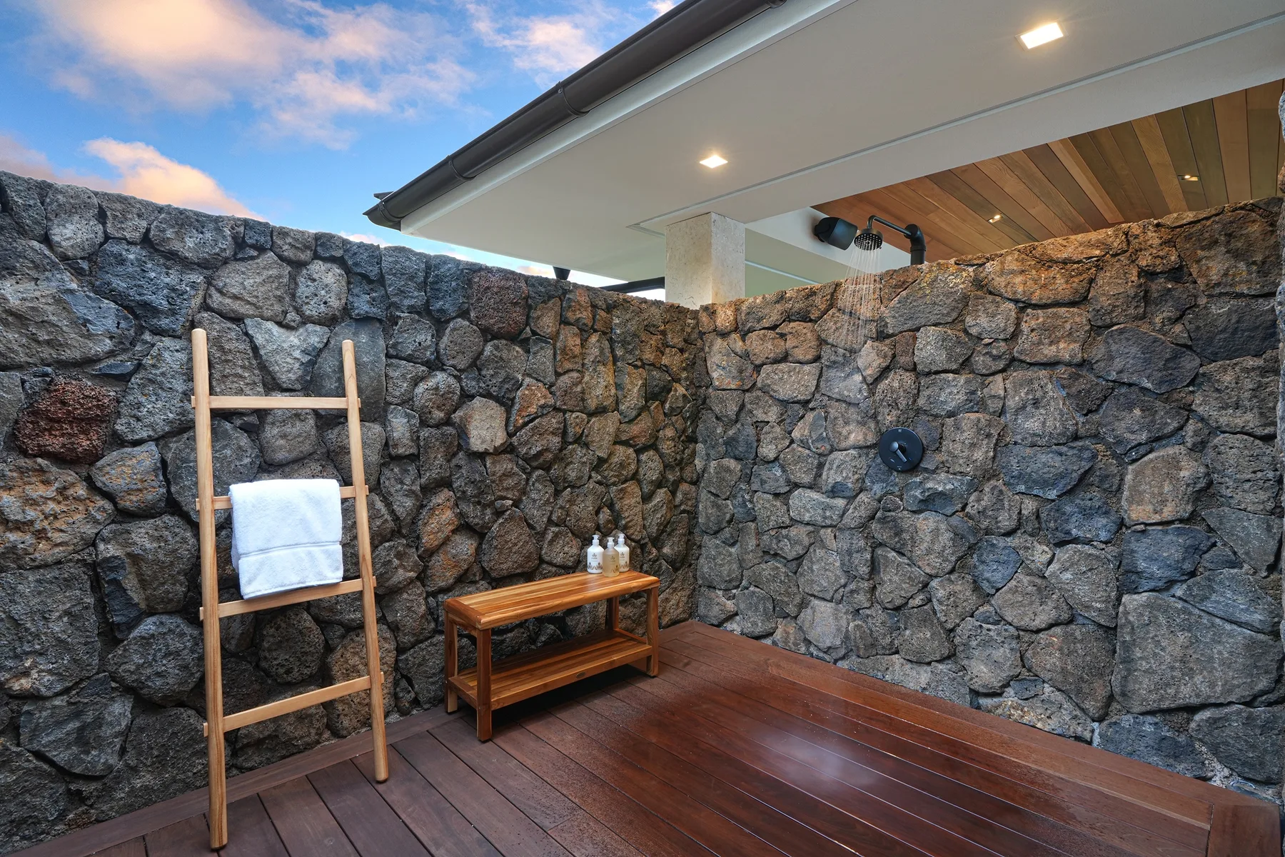 Una ducha exterior tiene paredes de piedra, una tarima de madera y un soporte para toallas.