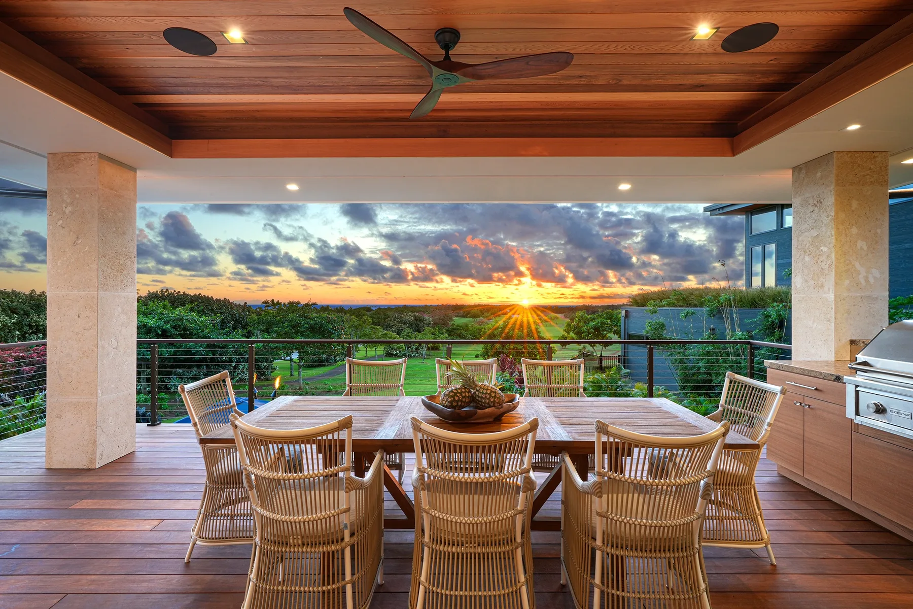 Una zona de comedor al aire libre da a una puesta de sol, con mesa y asientos bajo un techo revestido de madera.