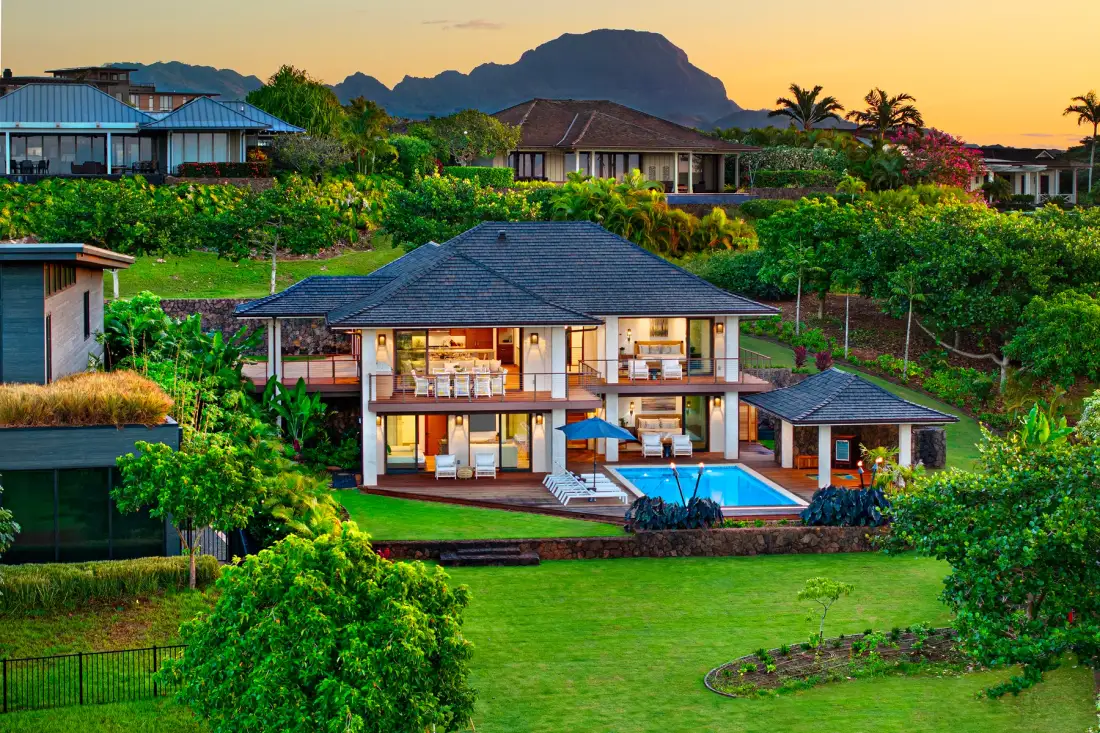 Una casa grande se alza junto a un jardín y una piscina, con montañas al fondo.