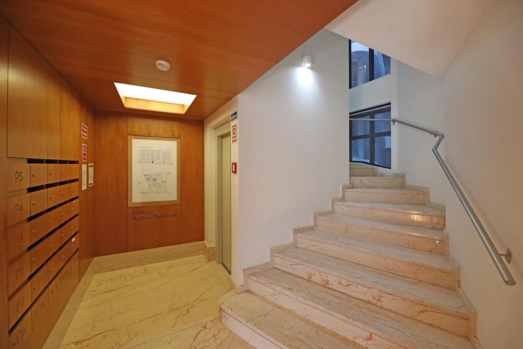 An indoor stairwell with marble steps, a metal handrail, and wooden mailboxes.