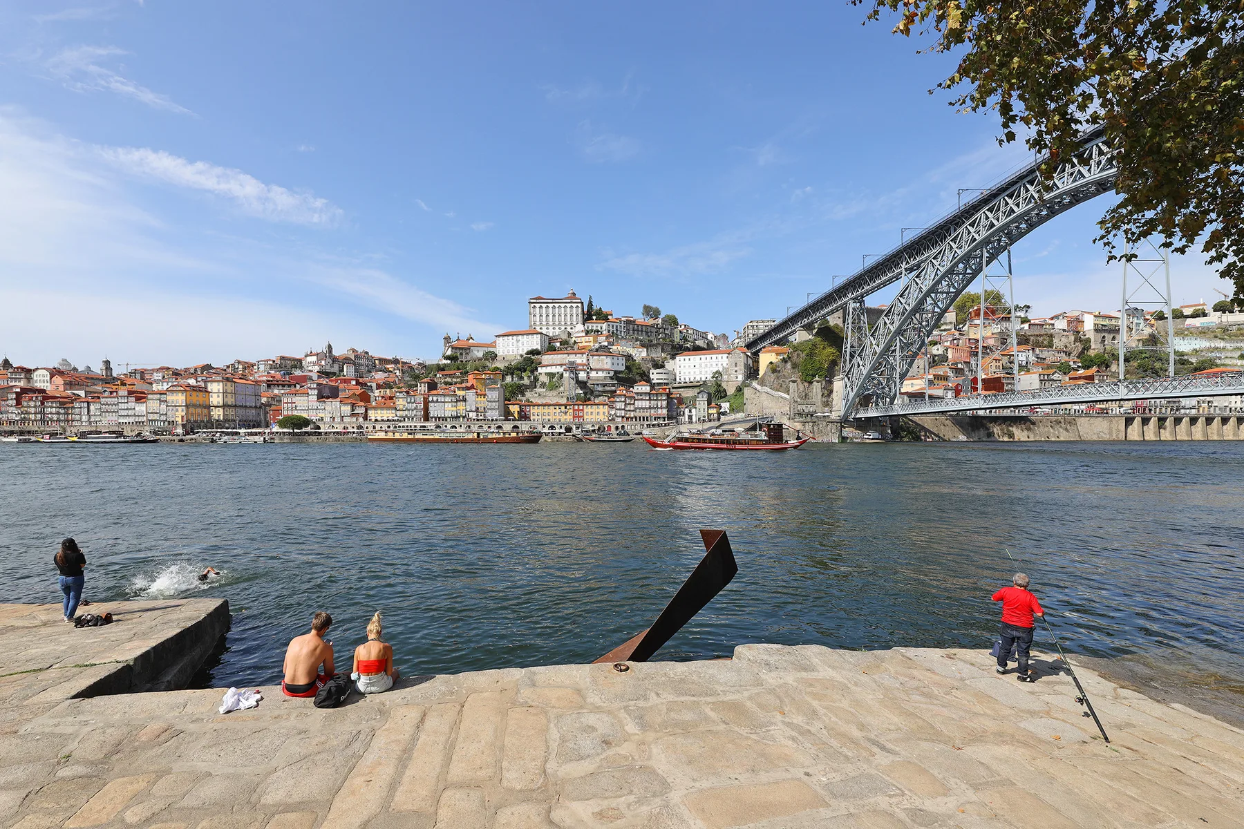 A riverside scene with people on a stone embankment and a bridge in the background.