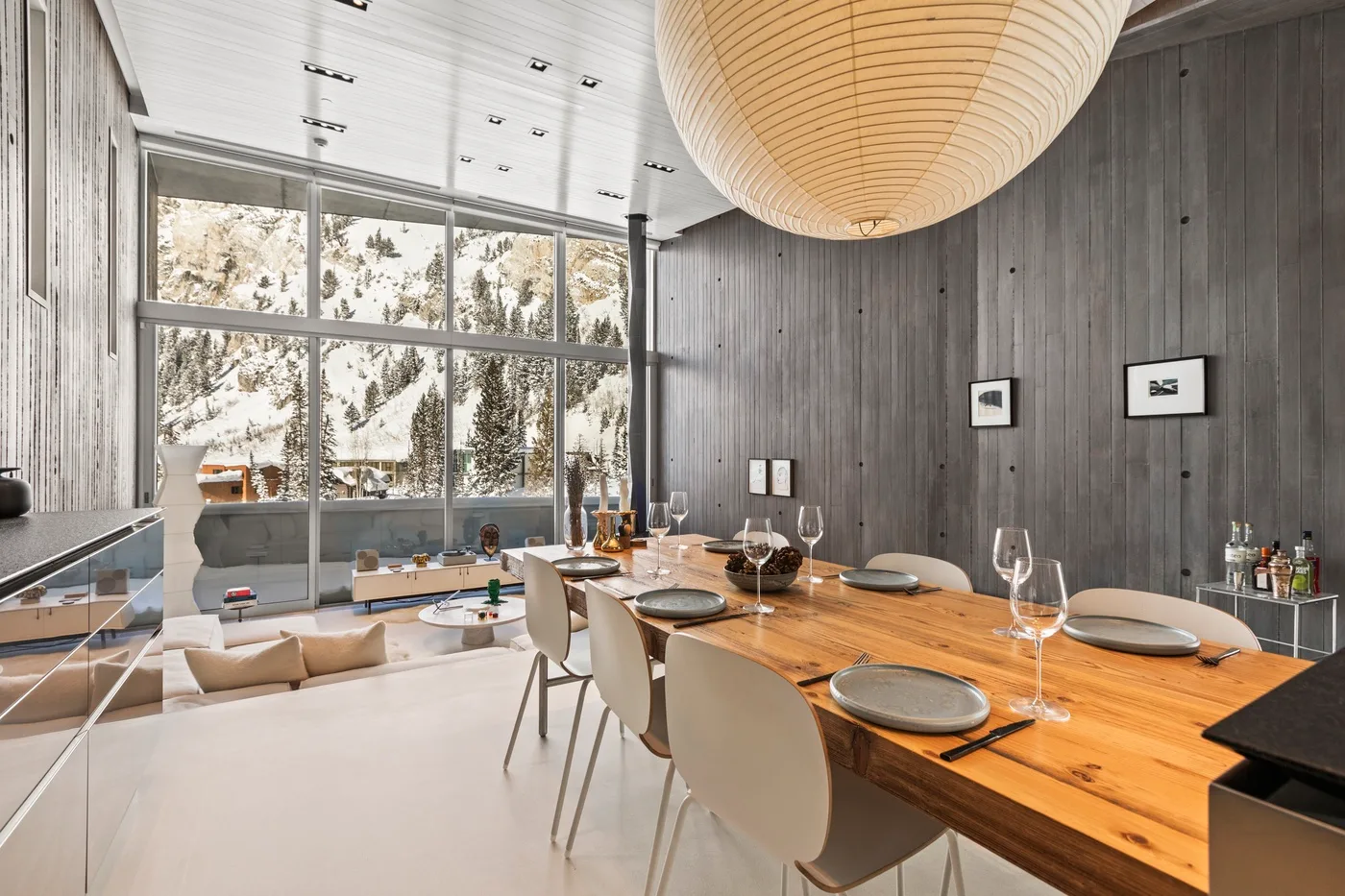 A dining area with a long wooden table beside large windows overlooking snowy trees.