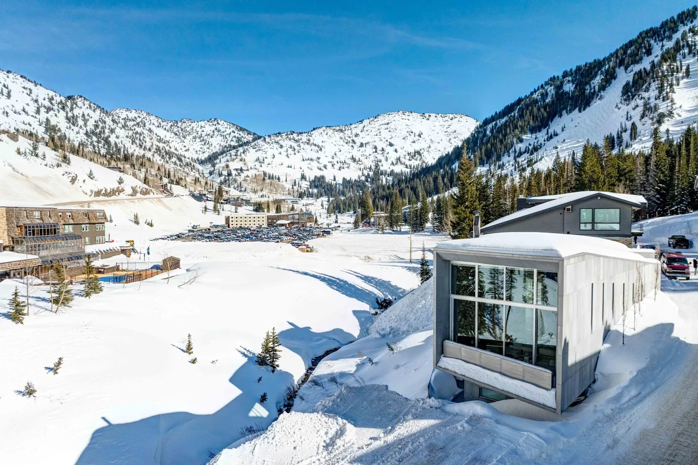 A modern building sits beside a snowy mountain valley under a clear blue sky.