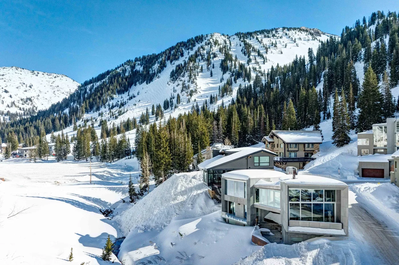Modern buildings sit beside a snowy mountain slope lined with evergreen trees.