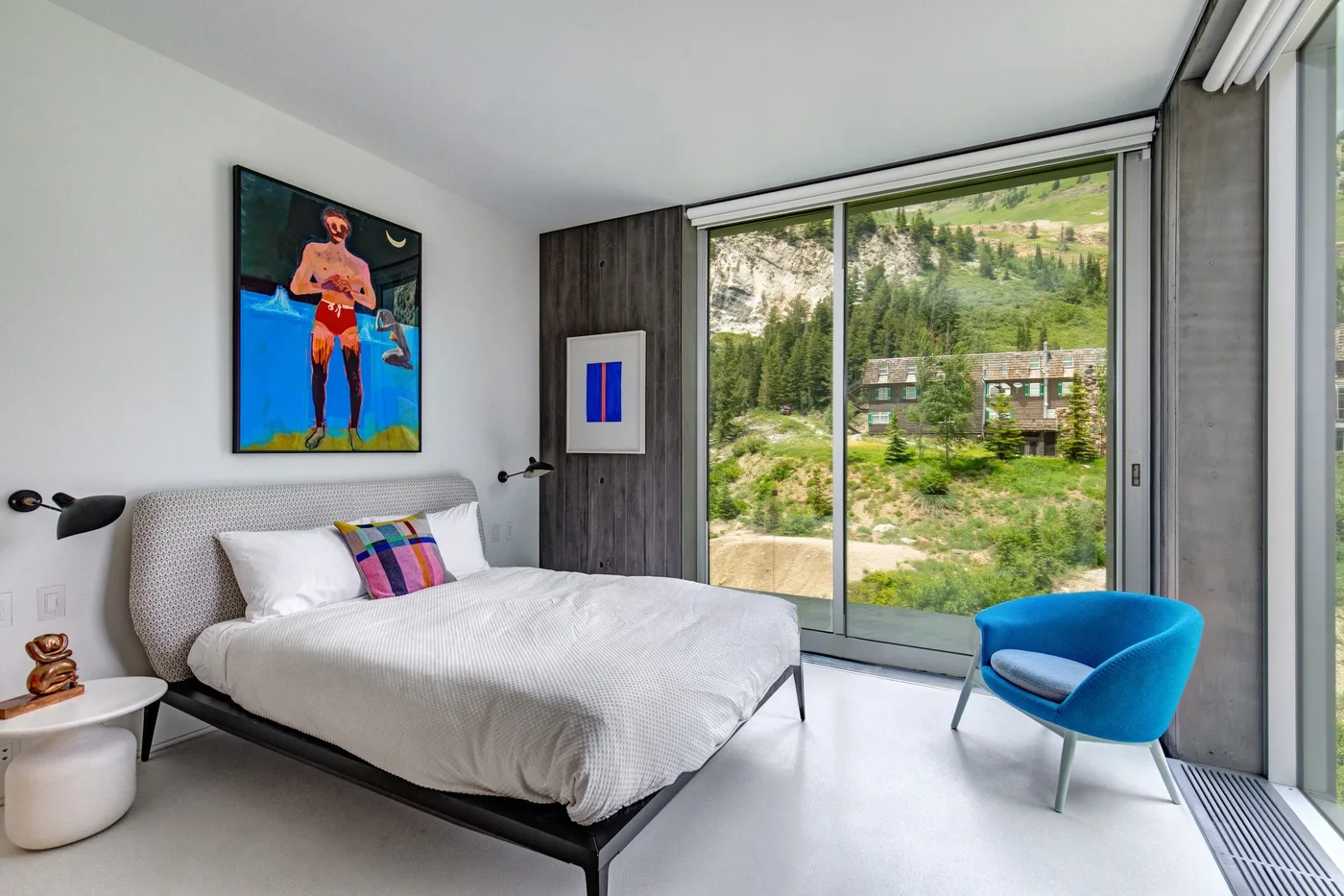 A bedroom with a bed and blue chair beside a large glass window overlooking greenery.