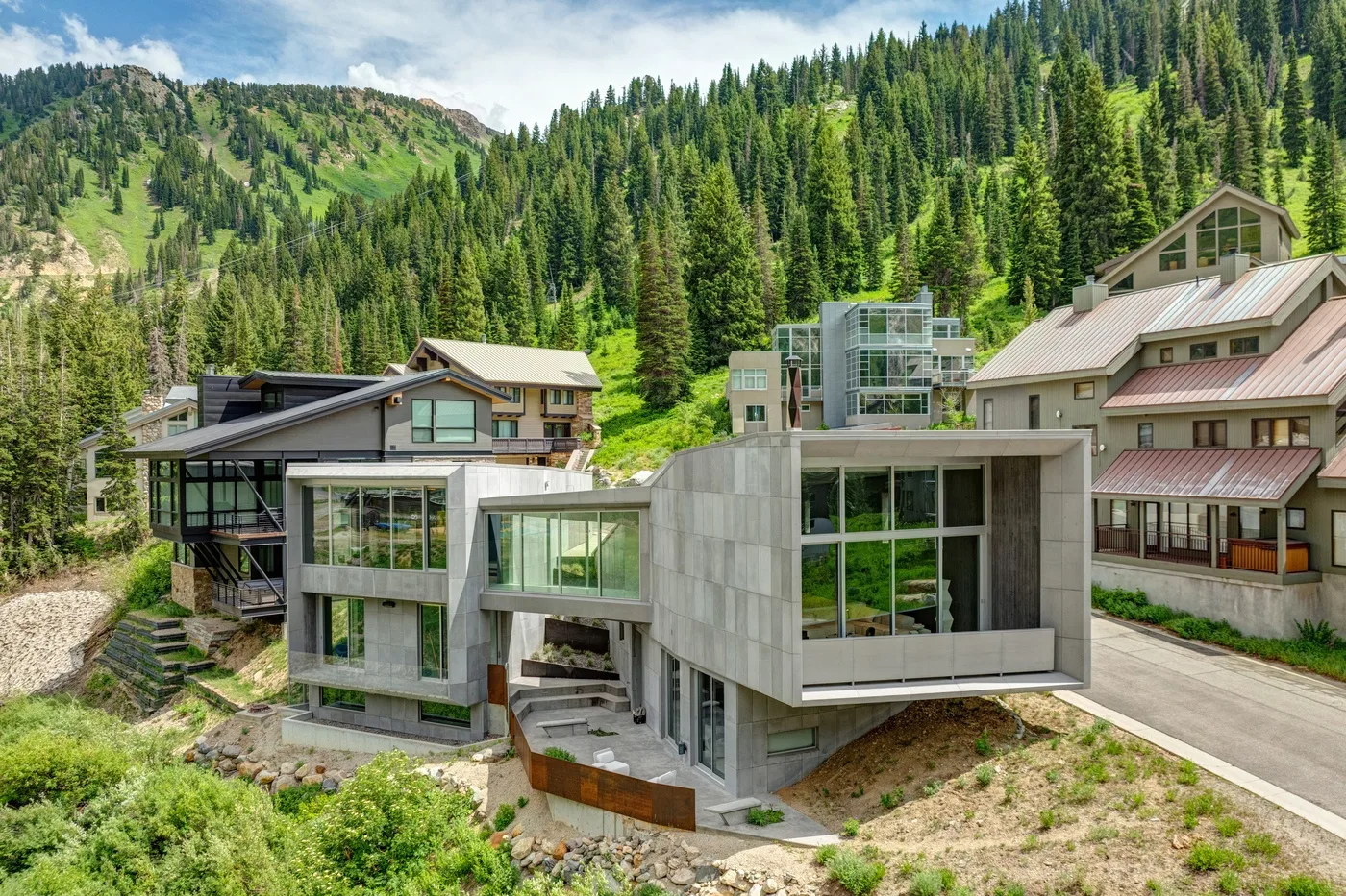 A modern house sits among wooded mountain slopes, with neighboring homes nearby.
