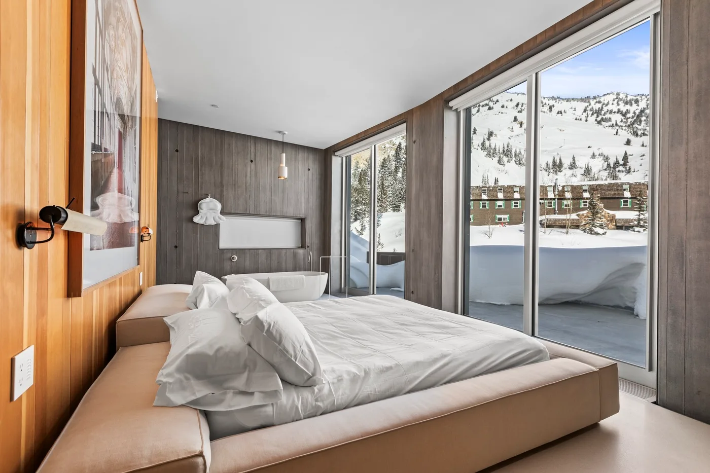 A modern bedroom with a bed beside large windows overlooking a snowy mountain scene.