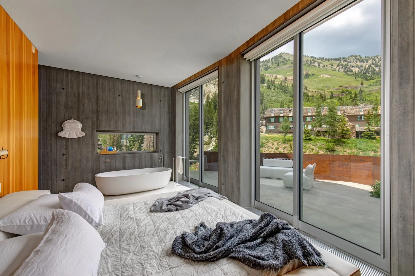 A bedroom with a bed and freestanding bathtub beside large windows overlooking a green hillside.