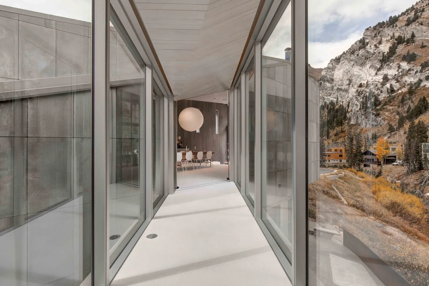 A glass-sided hallway leads toward a seating area, with mountain scenery visible outside.