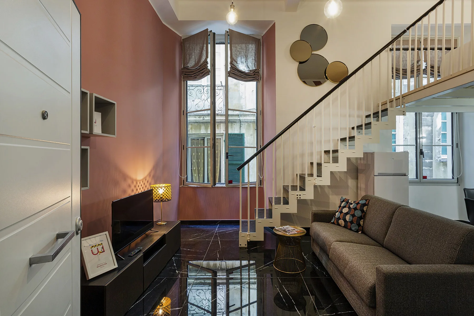 Una sala de estar con sofá, televisor y escalera junto a ventanas altas.