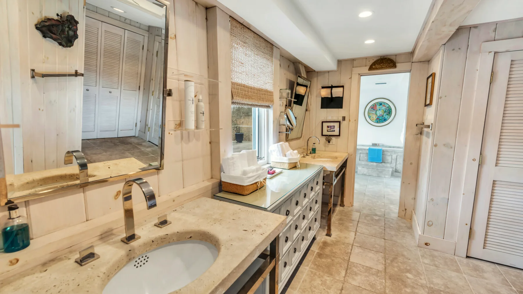 A bright bathroom with a long stone vanity, sinks, and a window.