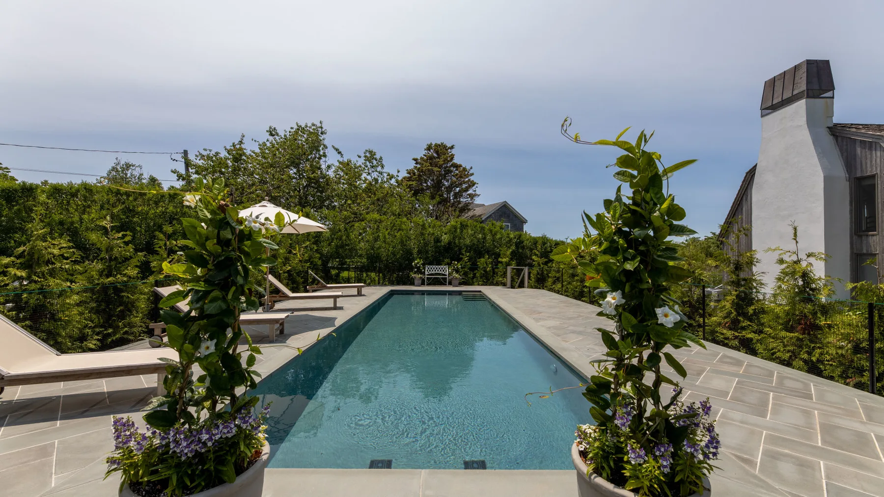 An outdoor swimming pool is surrounded by paved decking, greenery, and lounge seating.