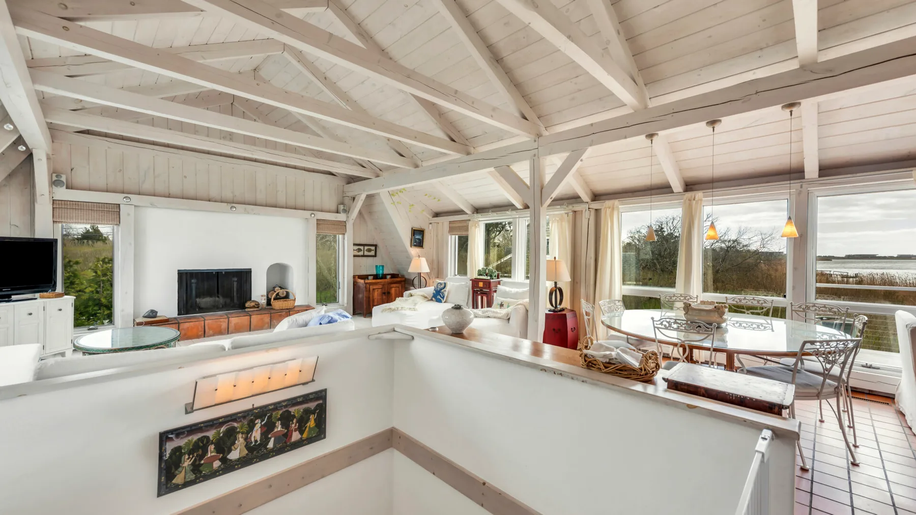 An open-plan living and dining area has white beams and large windows.