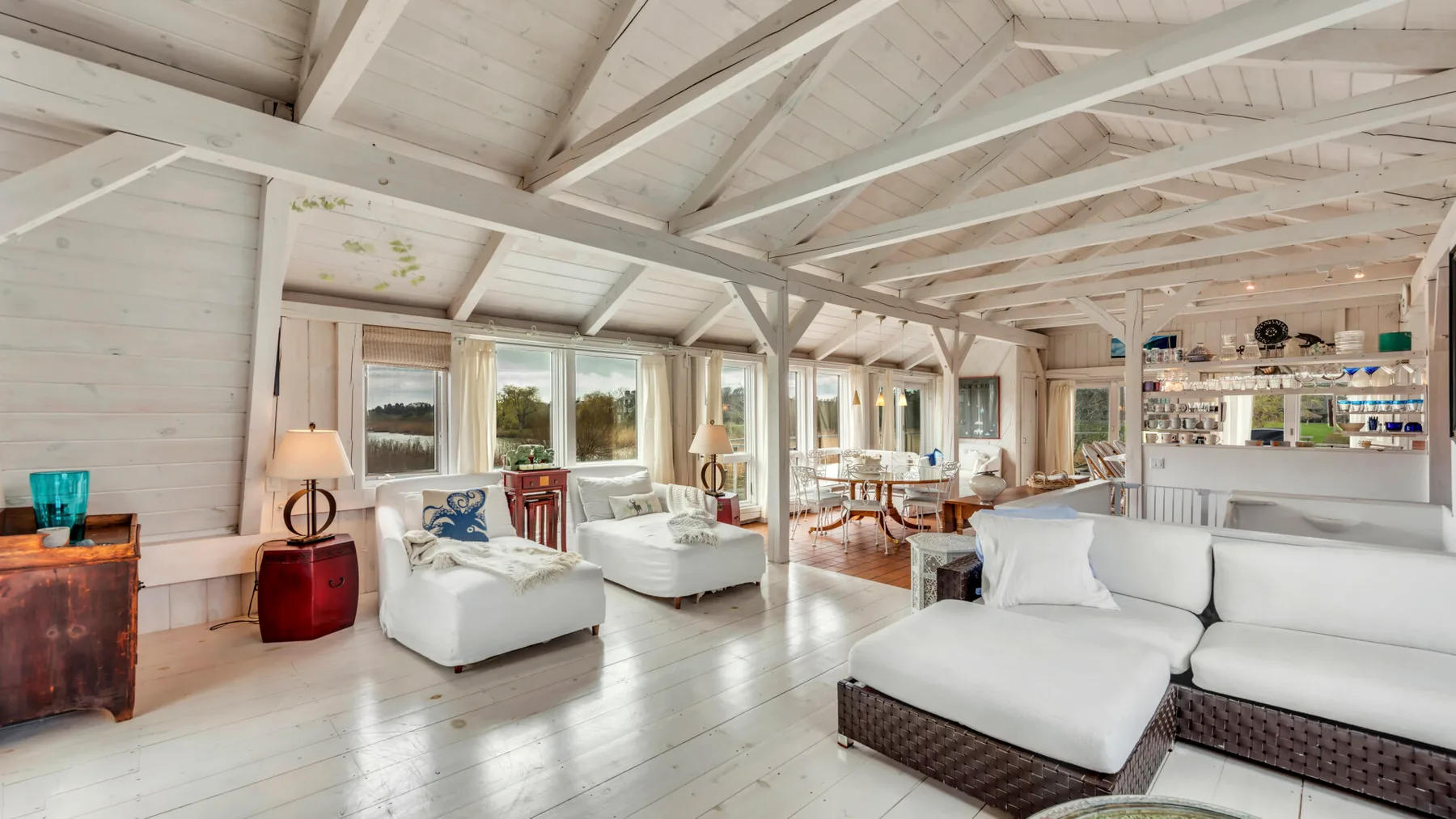 A bright living room with white seating beneath exposed beams and large windows.