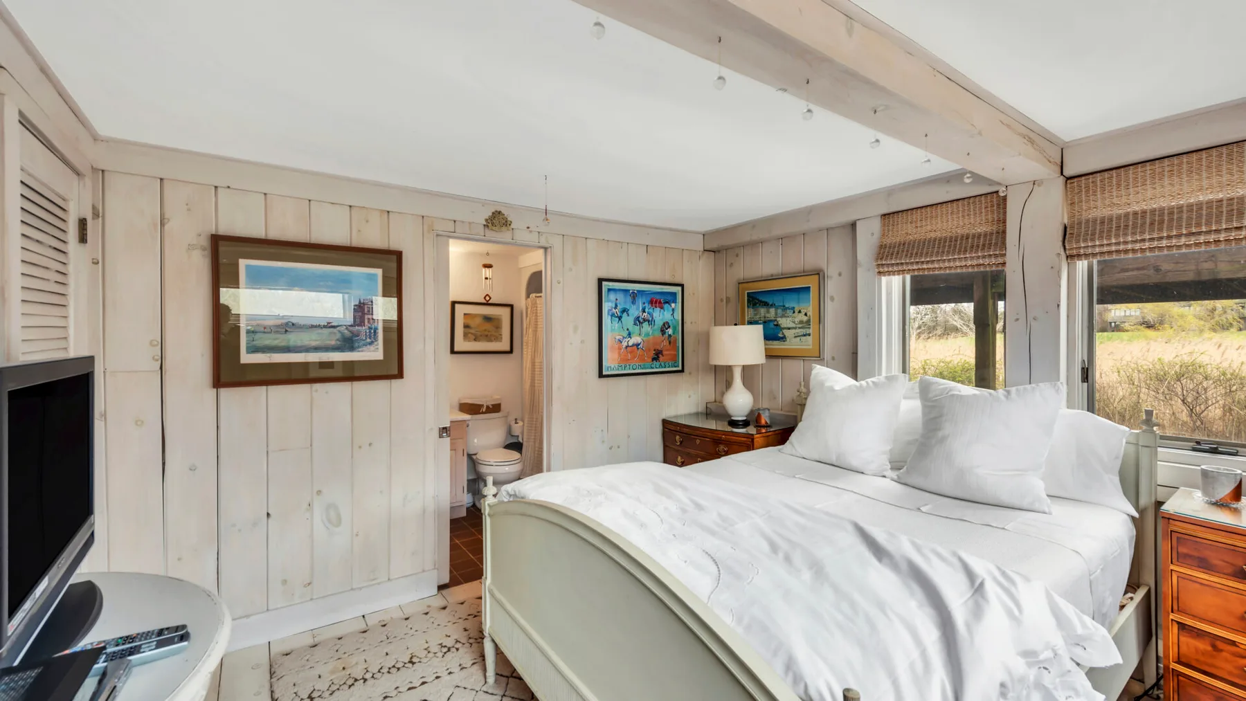 A bedroom with a large bed, framed artwork, and windows overlooking greenery.