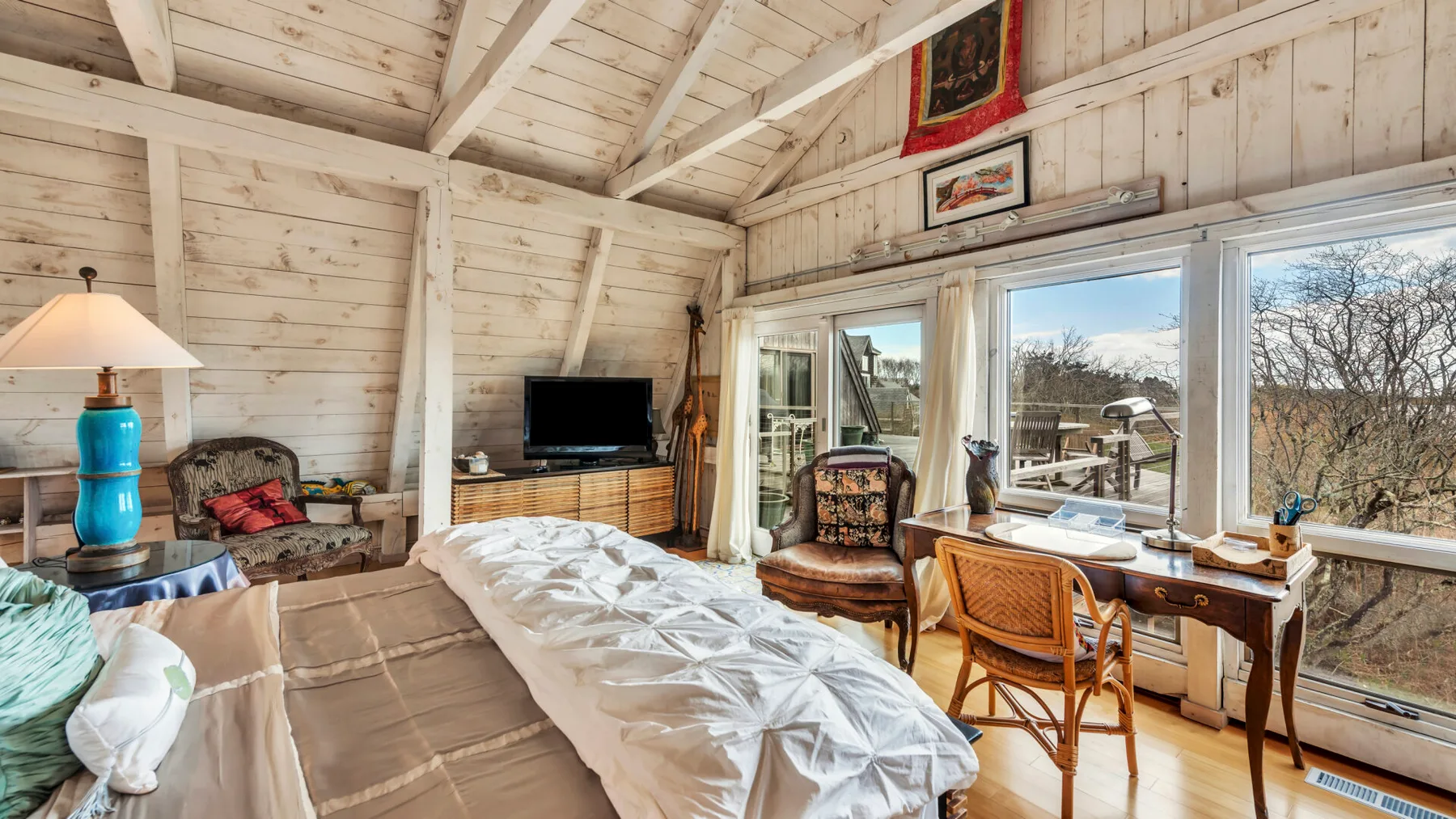 A bright bedroom with an unmade bed and large windows overlooking the outdoors.