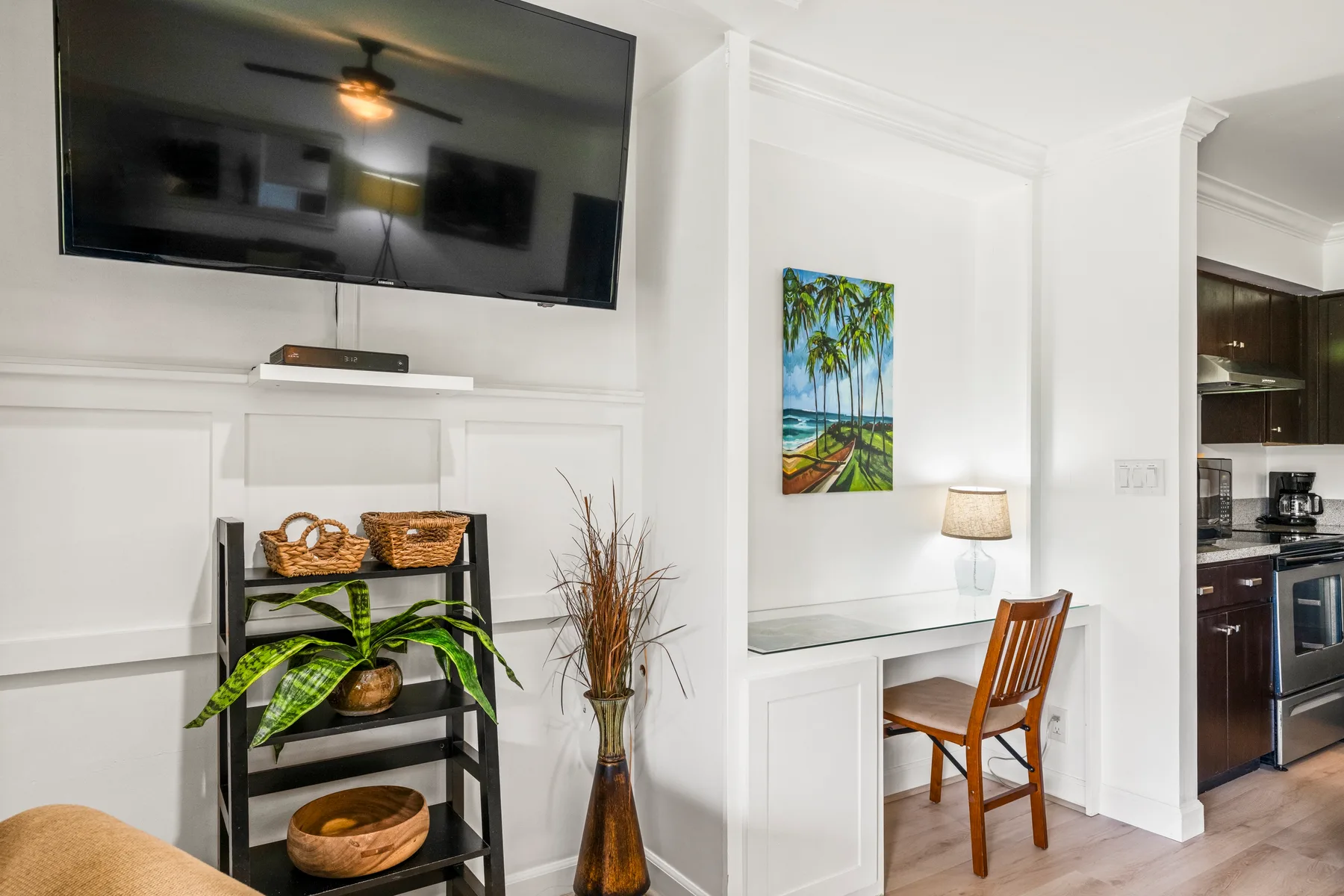 A bright living area with a wall-mounted television and a small desk beside the kitchen.