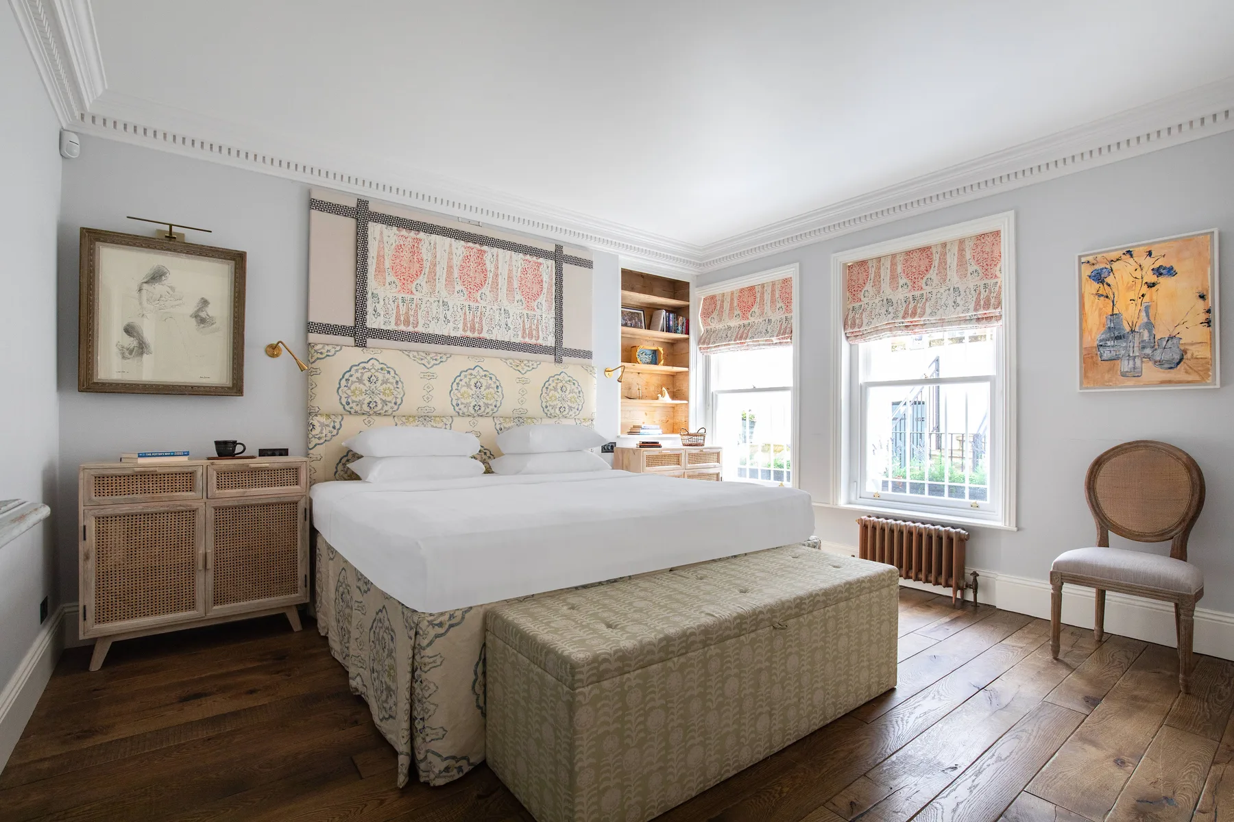 Un dormitorio luminoso con una cama grande, un banco tejido y ventanas altas.