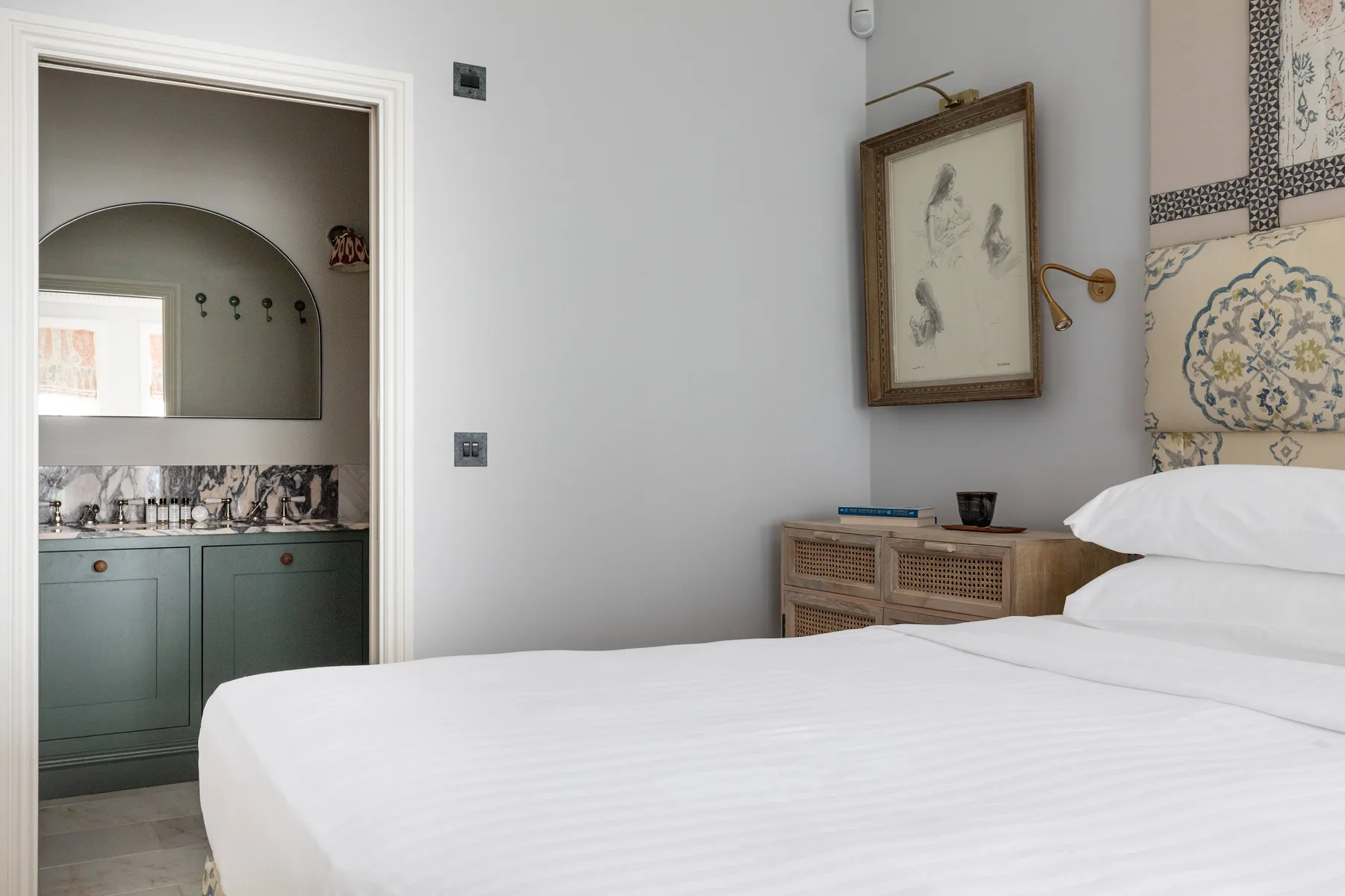 Un dormitorio con una cama blanca y un tocador visible a través de la puerta.