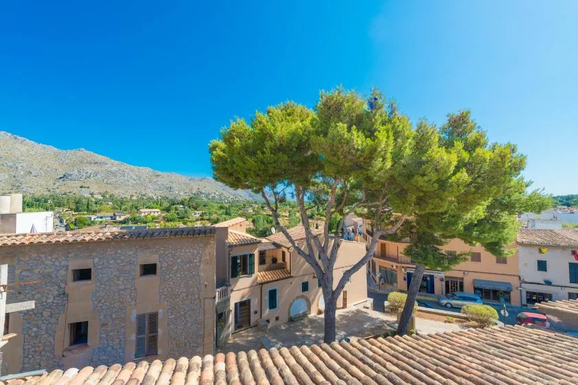 A village street with stone buildings, tiled roofs, and pine trees against a mountain backdrop.