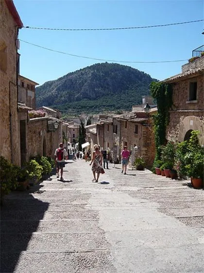 A sunlit village street runs between stone buildings, with a wooded mountain in the distance.