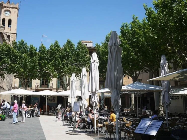 An outdoor café terrace with tables, umbrellas, and leafy trees.