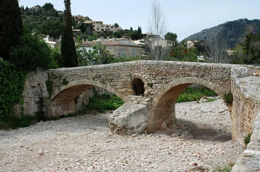 A stone bridge crosses a dry, rocky riverbed beneath a hillside.