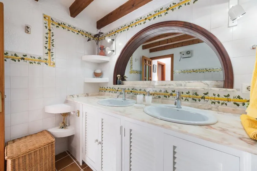 A bright bathroom has a long vanity with two sinks and a large arched mirror.