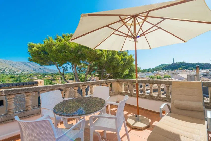 A sunny terrace has outdoor seating beneath a large umbrella, with hills in the distance.