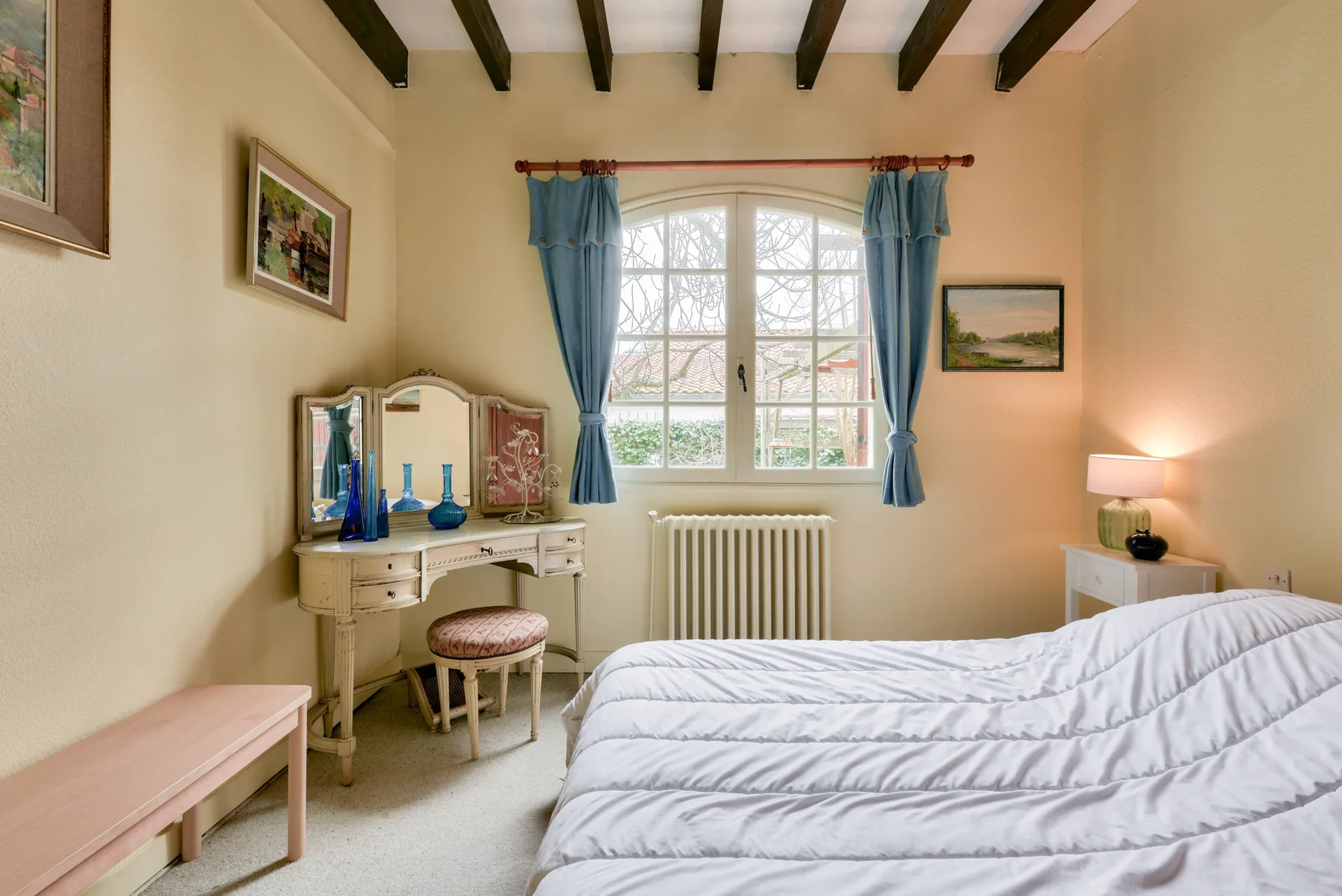 A bedroom with a bed, vanity, and a window framed by blue curtains.