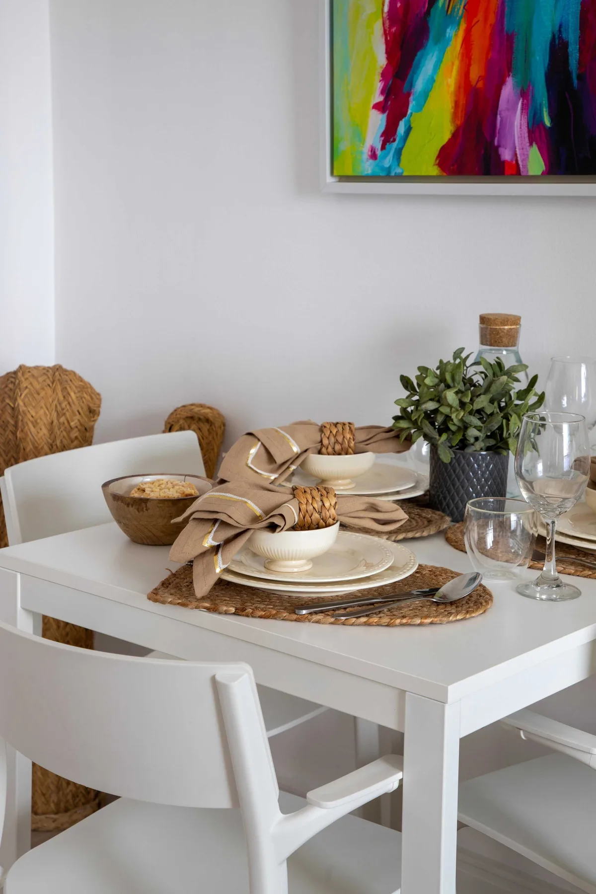 Una mesa de comedor está puesta con platos, servilletas dobladas, plantas y cristalería.