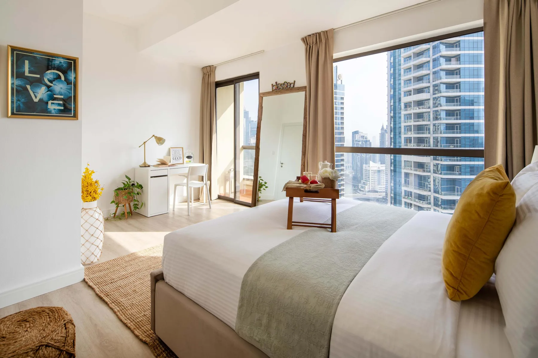 Un dormitorio con una cama bien tendida junto a grandes ventanales con vistas a edificios de la ciudad.