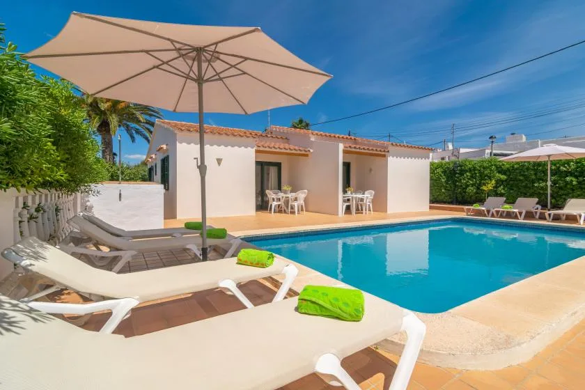 An outdoor swimming pool beside a white building, with loungers and a large shade umbrella.